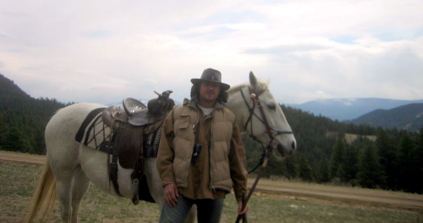 The guide in a cowboy hat beside a saddled white horse, mountains behind